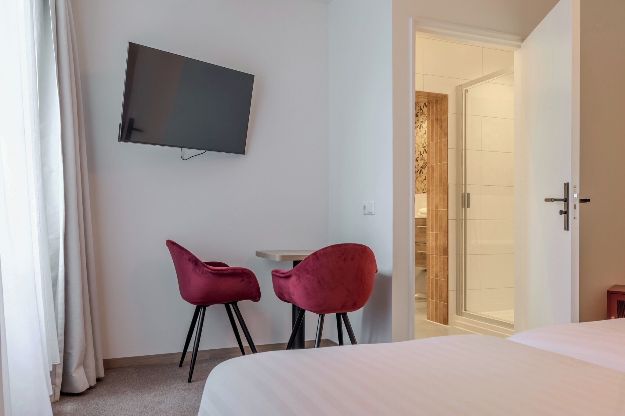 A hotel room with a bed, a TV mounted on the wall, two red chairs, and a small table.