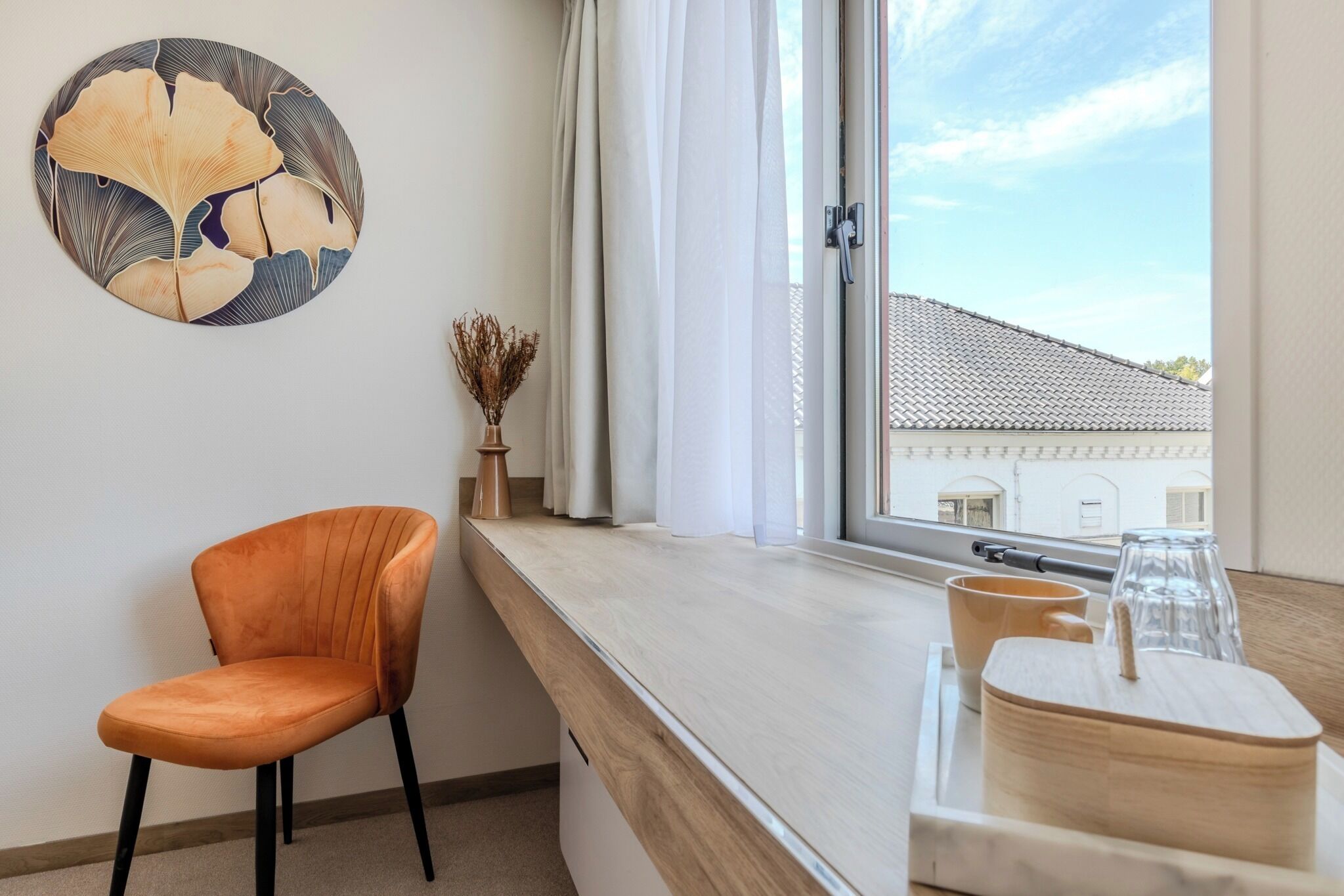 A room with a window, a chair, a table with cups and a vase, and a wall art piece.