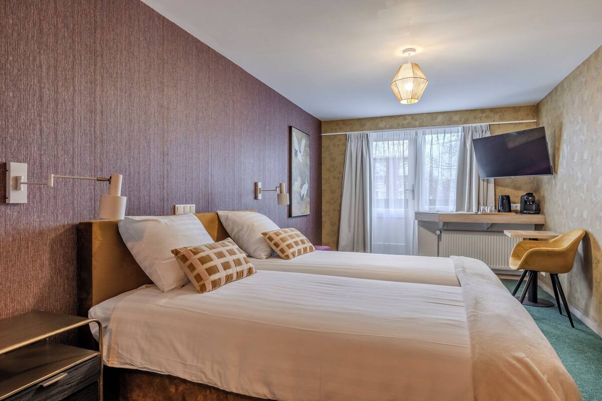Une chambre d’hôtel avec deux lits, un bureau avec une chaise, un téléviseur à écran plat et une fenêtre avec des rideaux.