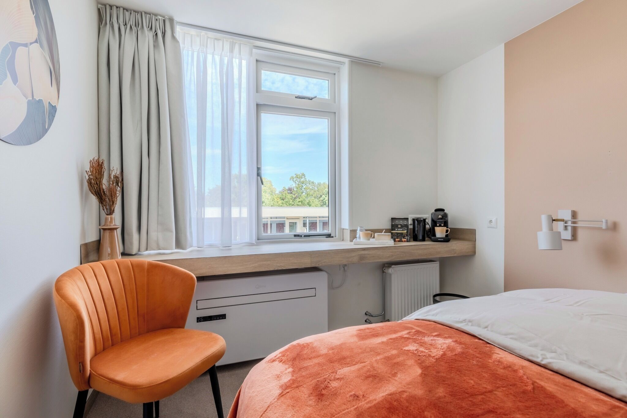 Habitación de hotel con cama, una silla, ventana con cortinas, radiador y cafetera sobre la encimera.