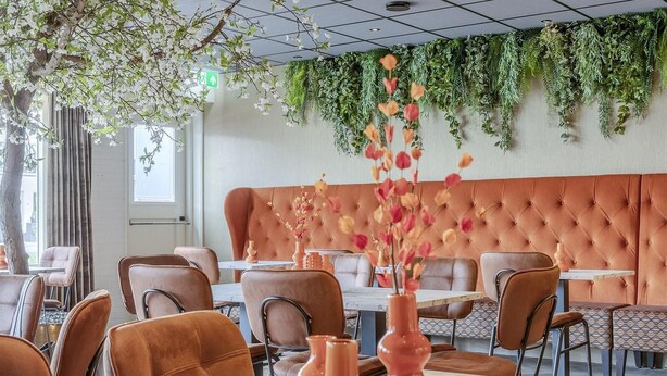 A restaurant with a tufted orange wall, brown leather chairs, and a tree with white flowers. - Hotel de Zon anno 1870 (Oosterwolde)
