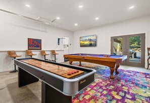 A pool table, foosball table, and bar stools in a room with a colorful carpet and a painting on the wall.