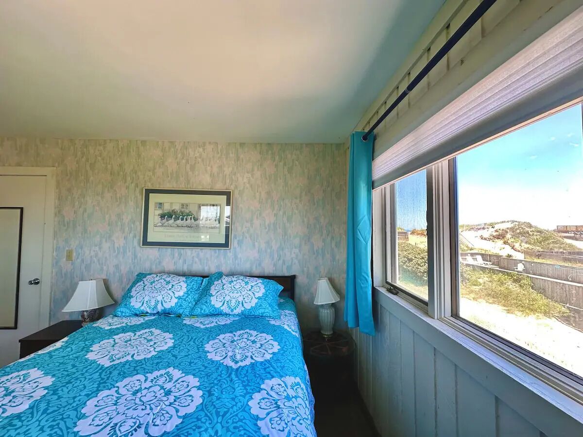 Una camera da letto con un letto, due lampade ai lati, un quadro appeso alla parete e una finestra con vista su una spiaggia e un ponte.