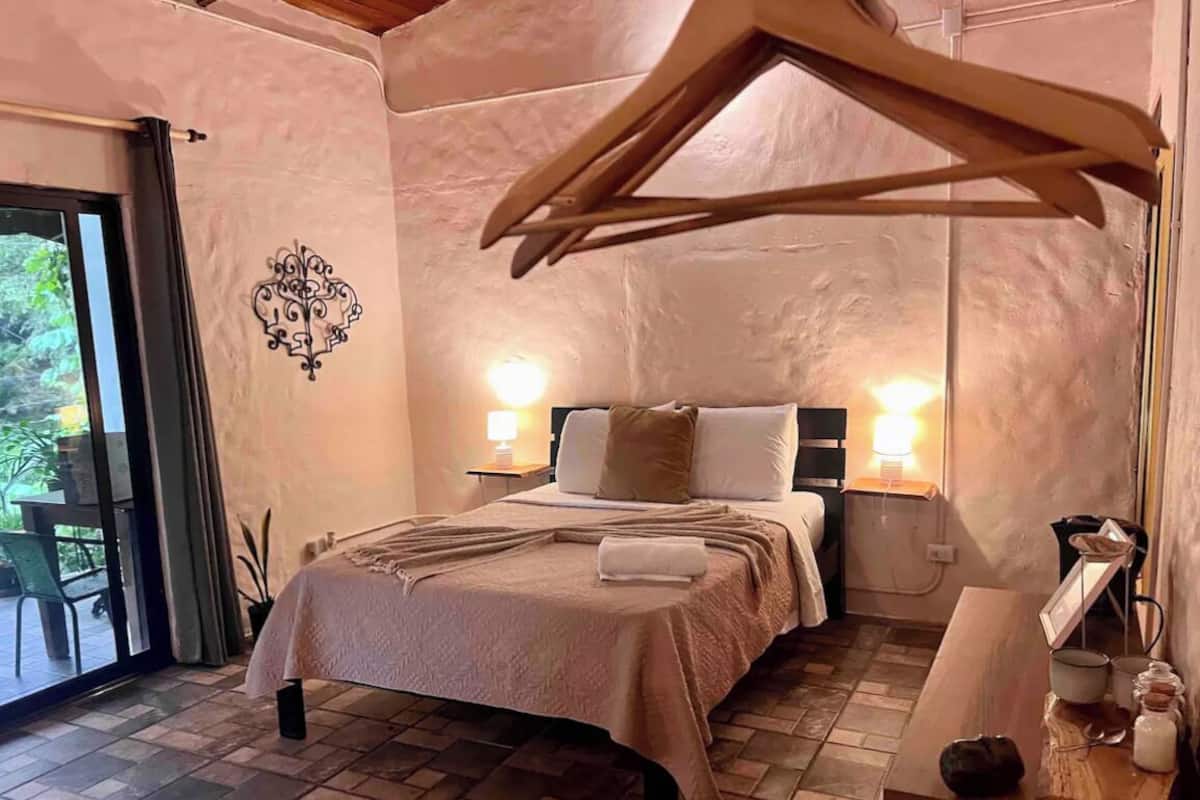 Quarto com cama, luminárias de cabeceira, uma mesa de madeira com xícaras e uma cadeira suspensa.