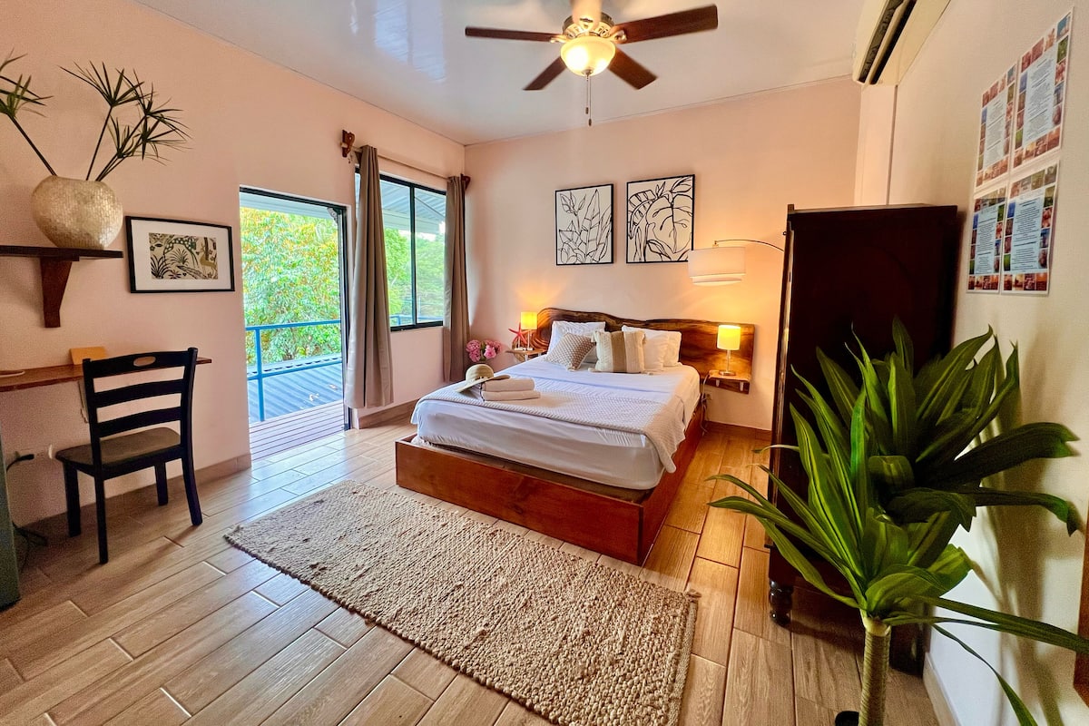 Quarto com cama, escrivaninha, cadeira, ventilador de teto e uma janela grande com cortinas.