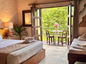 A bedroom with a bed, a bedside table with a lamp, a potted plant, and a view of a balcony with a table and chairs. - Kikazaru Boutique Hotel (Quepos)