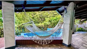 A hammock under a covered area with a view of a swimming pool and lush greenery. - Kikazaru Boutique Hotel (Quepos)