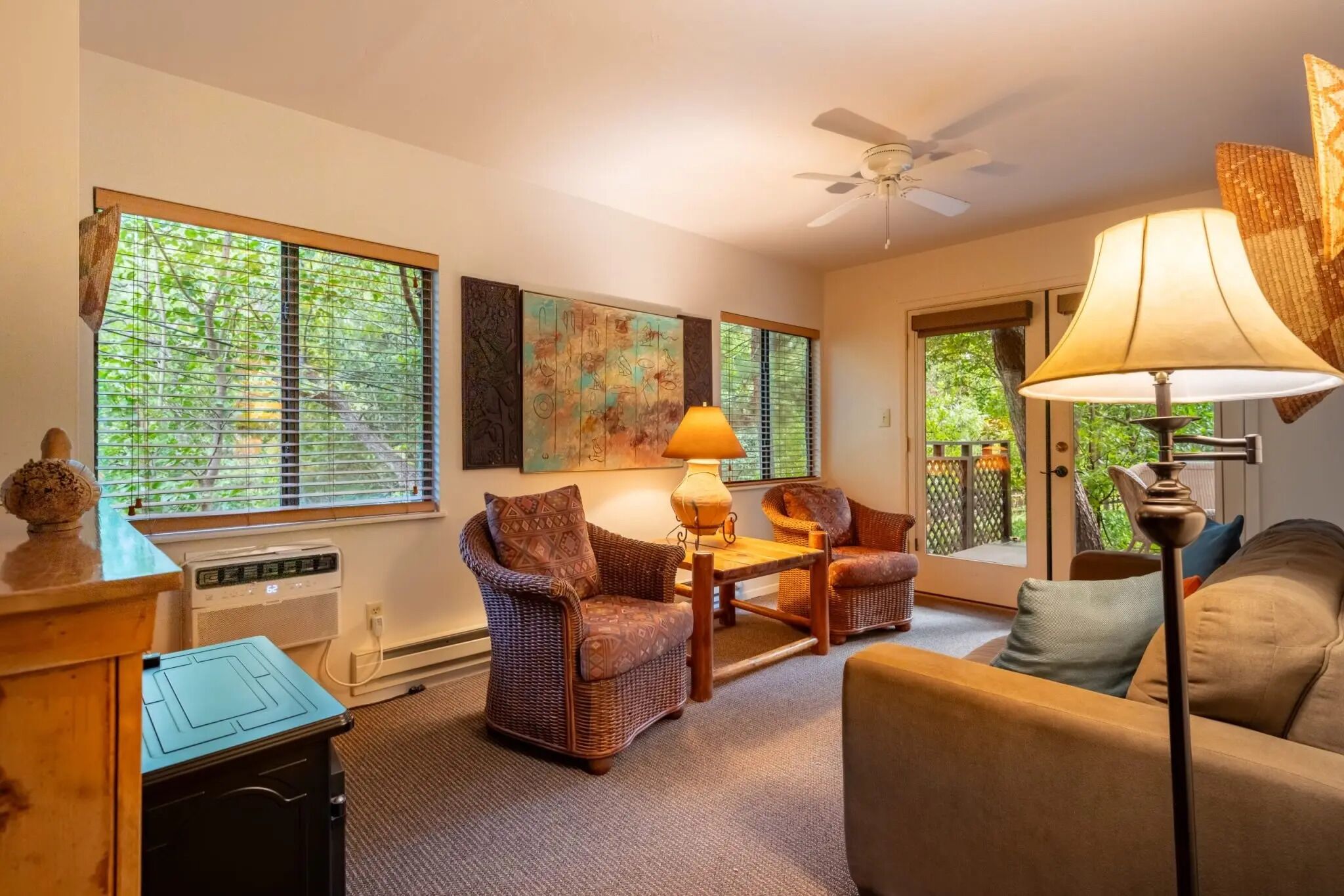 A cozy living room with wicker furniture, a ceiling fan, and large windows with blinds.