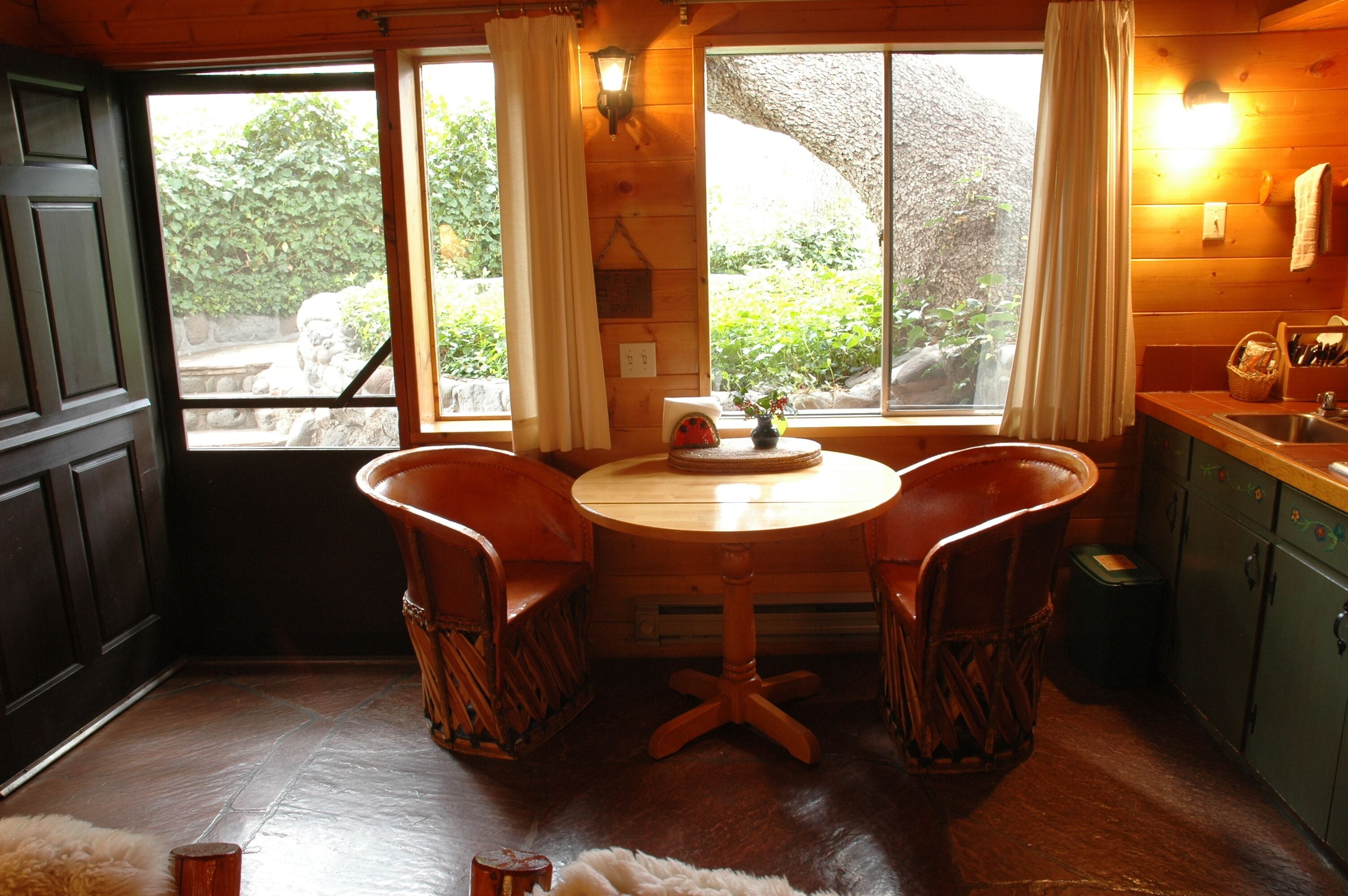A cozy wooden cabin interior with a dining table and chairs, a window with curtains, and a kitchen area with cabinets and a sink.