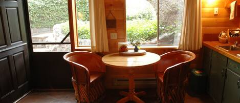 A cozy wooden cabin interior with a dining table and chairs, a window with curtains, and a kitchen area with cabinets and a sink.