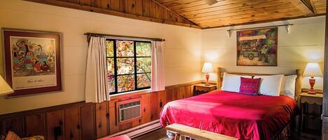 A cozy room with a wooden ceiling, a large bed with red bedding, a window with curtains, and a painting on the wall.