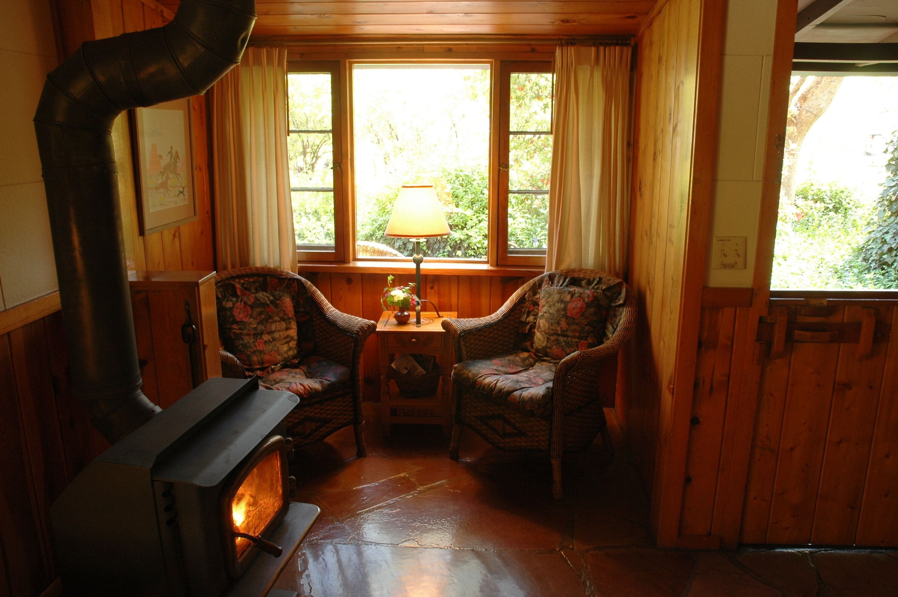 A cozy room with a wood-burning stove, two wicker chairs, a small table with a lamp, and a window with curtains.
