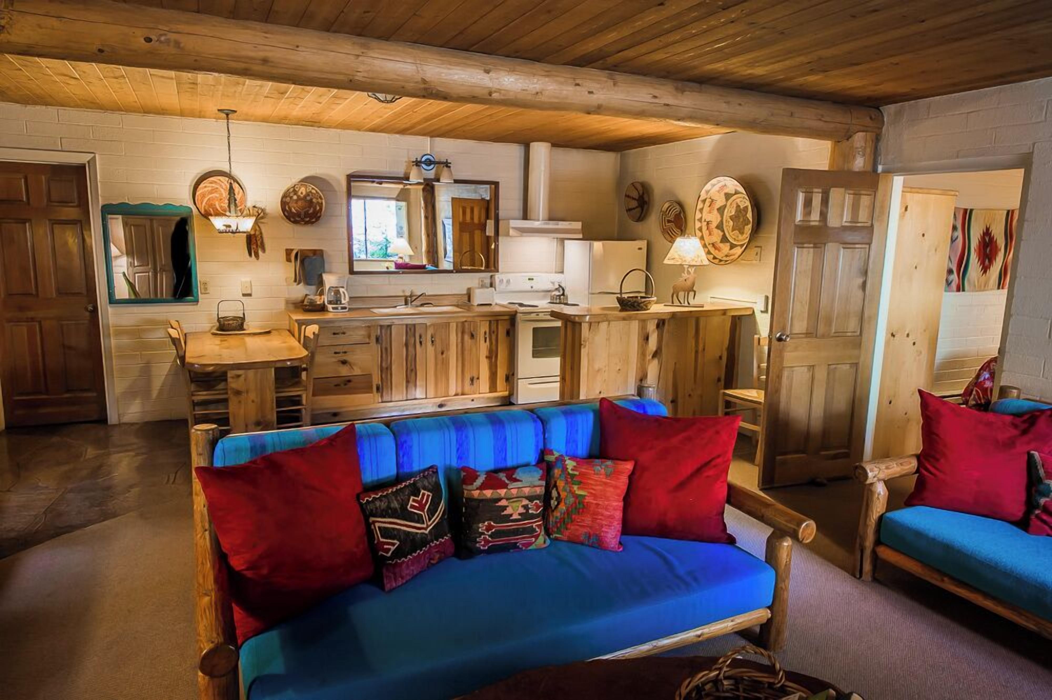 A rustic cabin interior with a wooden dining table, blue sofa, and a kitchen area with a stove and sink.