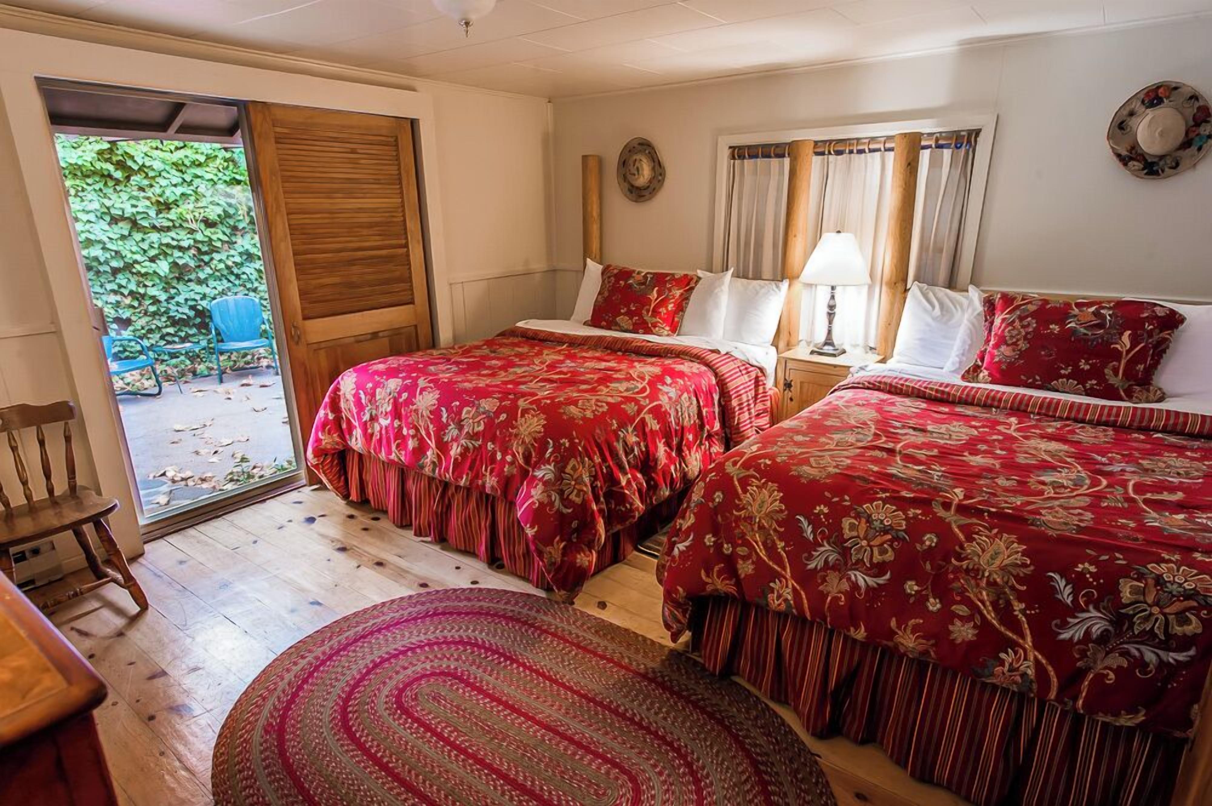 Quarto com duas camas, piso de madeira, porta que dá para o exterior e uma pequena mesa com abajur.