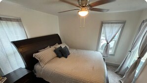 Un dormitorio con cama, mesita de noche, ventilador de techo y ventanas con cortinas.