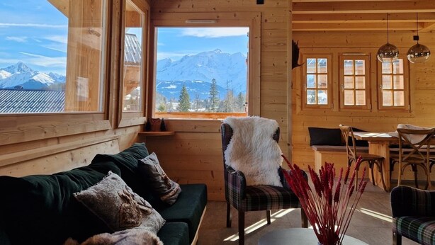 Een knus houten vakantiehuisje met een bank, een stoel en een eettafel. Een groot raam biedt uitzicht op bergen en bomen.