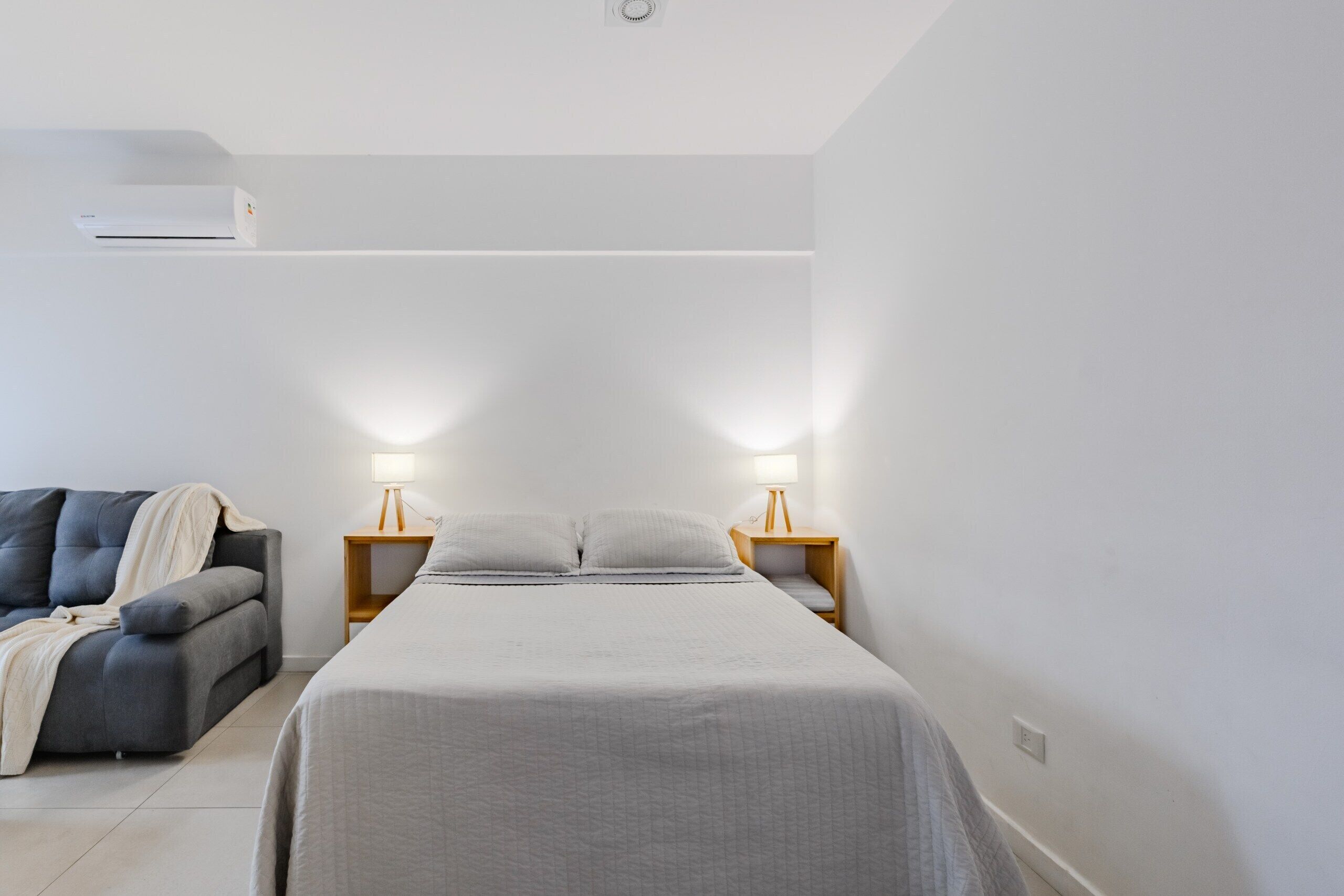 A minimalist bedroom with a bed, bedside lamps, a sofa, and an air conditioner.