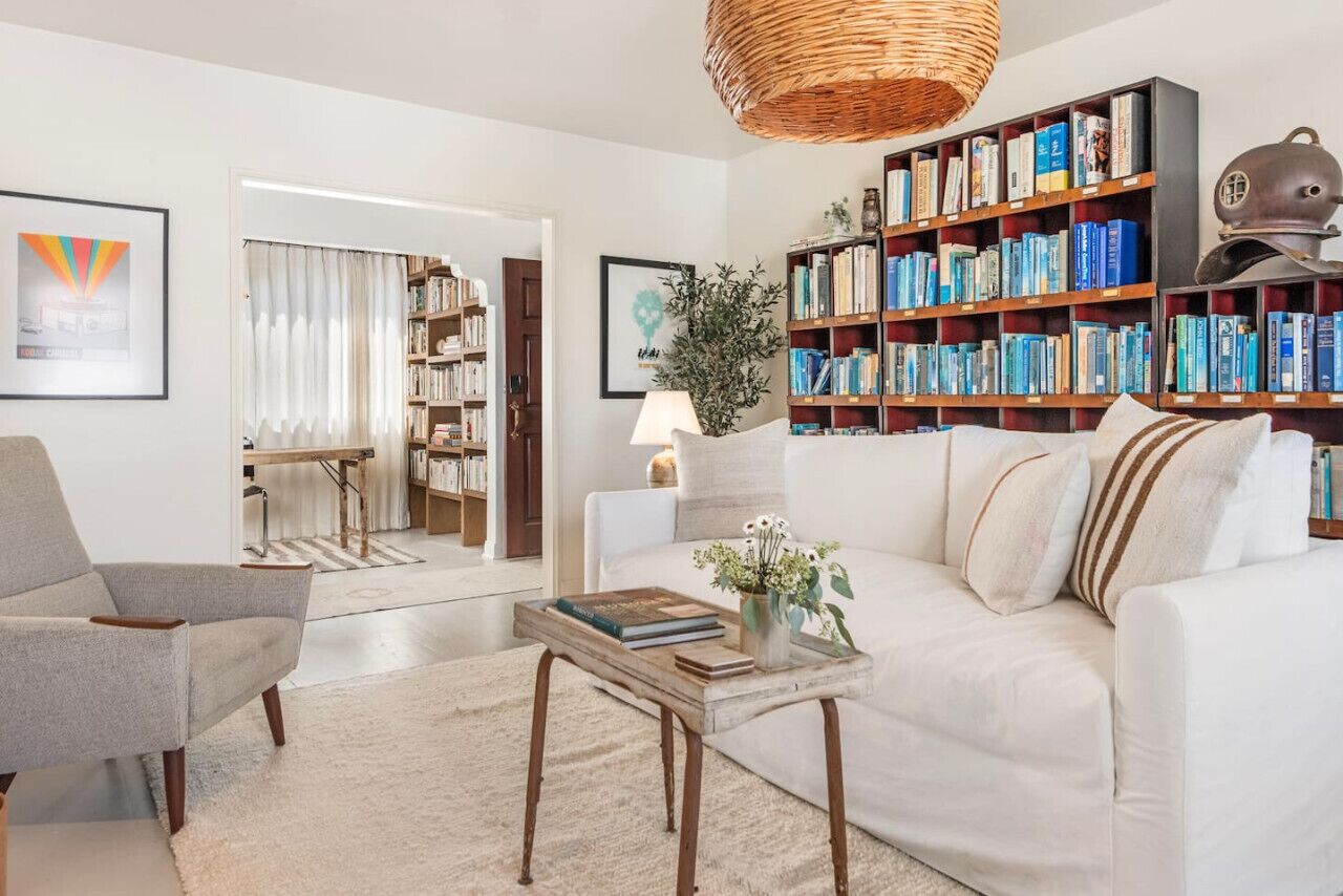 Un salon avec un canapé blanc, un fauteuil gris, une table basse en bois, une bibliothèque et un luminaire suspendu en osier.