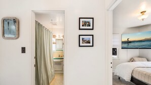 A hotel room with a bed, a large window with a view of the sea, a bathroom with a shower curtain, and two framed pictures on the wall.