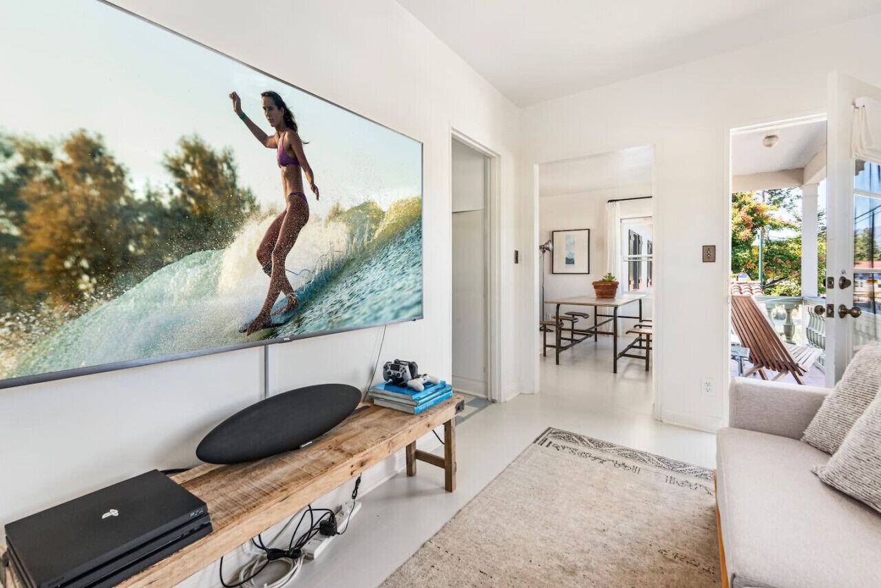A modern living room with a large TV displaying a surfing scene, a wooden bench with a laptop, and a dining area in the background.