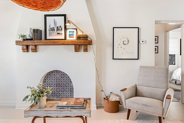 A cozy living room with a fireplace, a wooden shelf with books and decorative items, a comfortable armchair, and a wall-mounted picture.