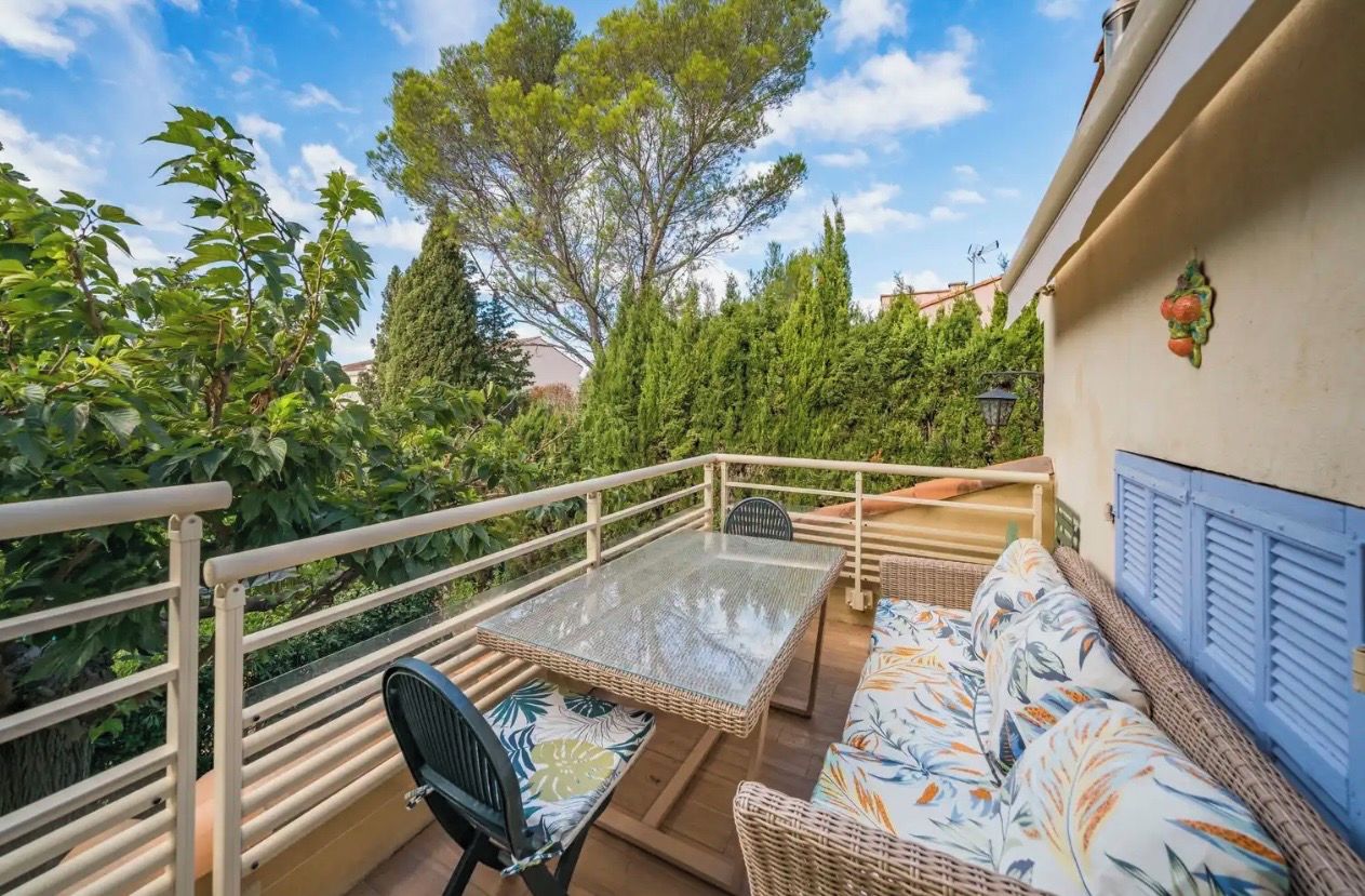 Een balkon met een rieten tafeltje en stoelen, een bank met bloemenkussens en een leuning.