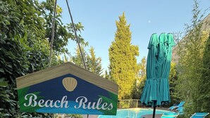 Una terrassa de fusta amb un cartell que diu 'Beach Rules', una piscina amb gandules i un parasol verd.