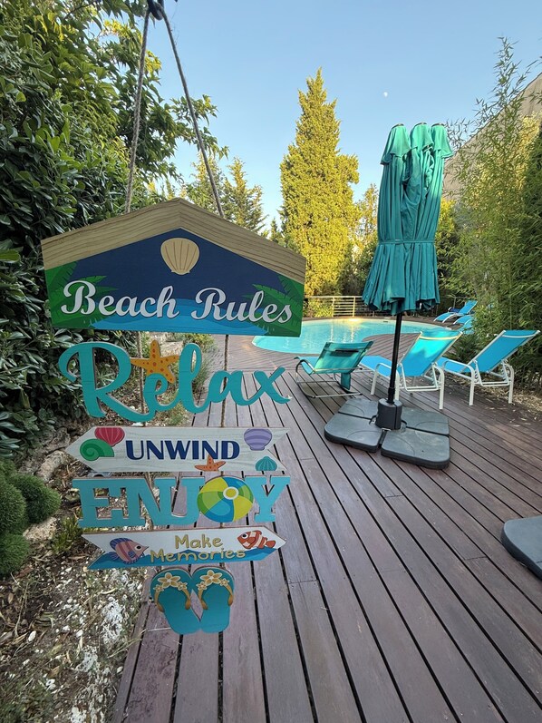 A wooden deck with a sign reading 'Beach Rules,' a pool with lounge chairs, and a green umbrella.