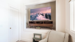 Un salon avec un téléviseur fixé au mur qui diffuse une scène de montagne, un meuble de rangement en bois, un canapé à carreaux et un petit tabouret vert.
