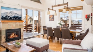 Un salon chaleureux avec une cheminée, des sièges confortables, un coin repas et de grandes fenêtres offrant une vue sur les montagnes.