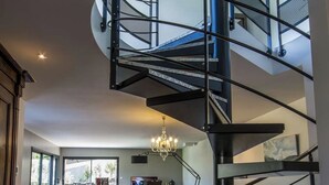 Un interior moderno con una escalera curva que lleva a un entrepiso, un comedor con candelabro y vistas al exterior.