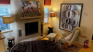 A living room with a fireplace, a large abstract painting, a comfortable armchair, and a small table with a lamp.