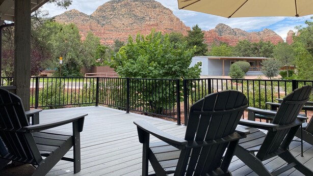 A wooden deck with Adirondack chairs and a large umbrella, overlooking a mountainous landscape with a small building in the background.