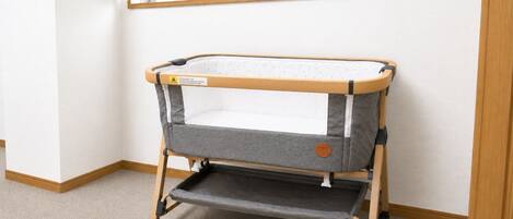 A portable crib with wheels, a padded mattress, and a lower shelf, set against a plain wall with a window above.