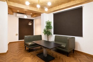 A room with a wooden herringbone floor, a dark brown table, two green upholstered sofas, a potted plant, and a large dark screen on the wall.