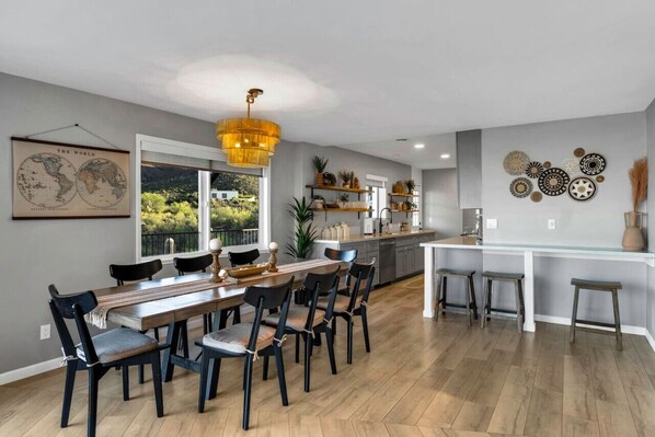 A modern kitchen with a dining area, featuring a large wooden table, black chairs, and a hanging light fixture.