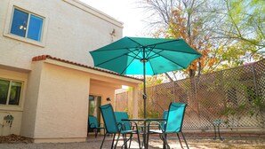 En terrasse med et bord og stole under en grøn parasol, en hvid bygning med et vindue, og et hegn med træer i baggrunden.