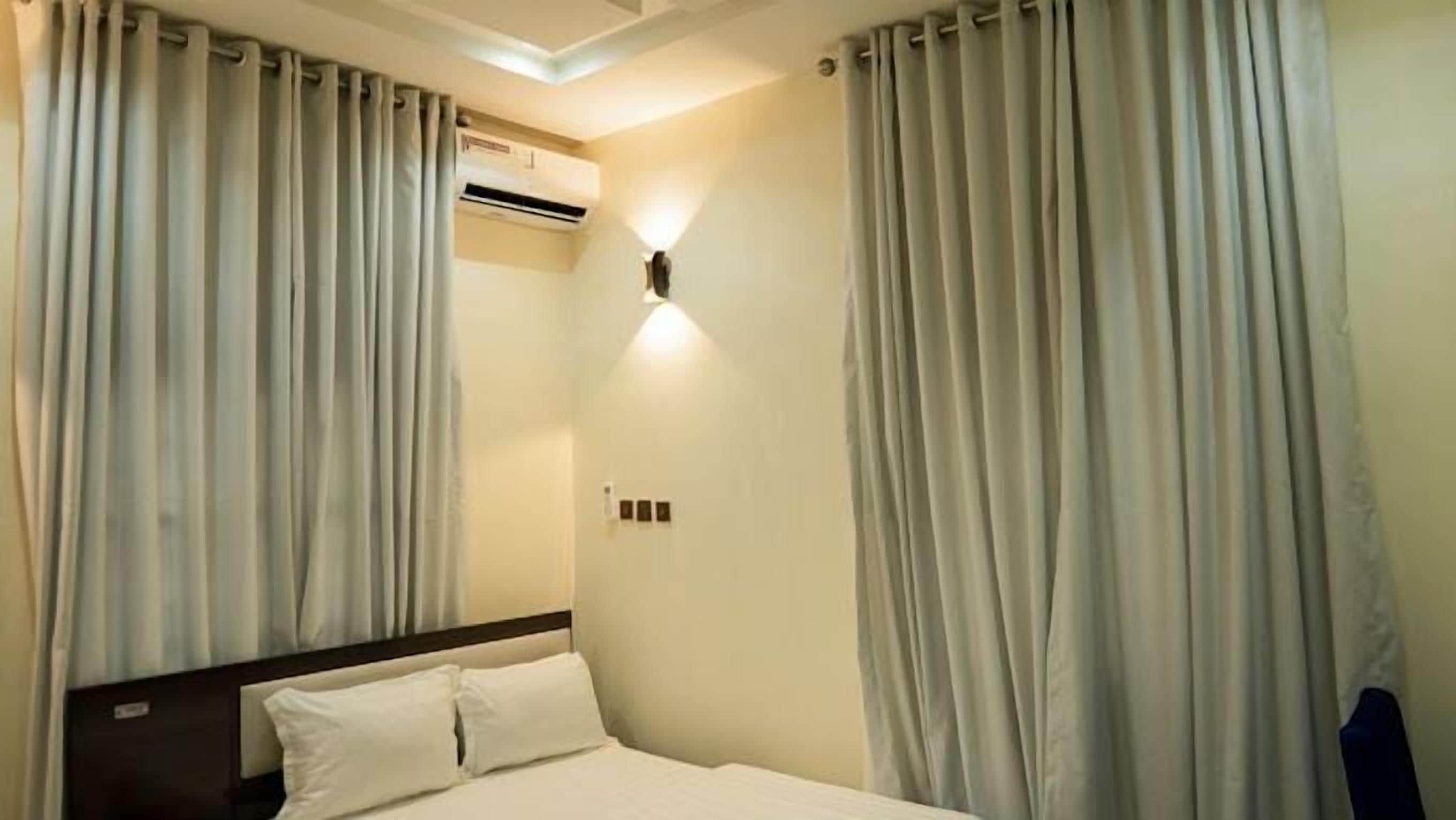 A hotel room with a bed, curtains, an air conditioning unit, and a wall-mounted light fixture.
