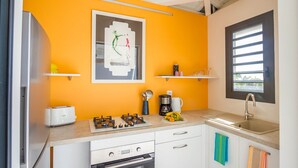 Une cuisine compacte équipée d’une cuisinière à gaz, d’un four et d’une machine à café posée sur le plan de travail. On y trouve deux placards blancs et une fenêtre avec des stores.