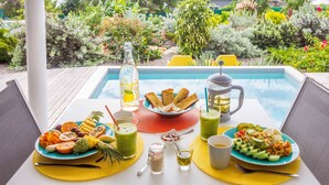 Une table à manger dressée avec une variété de plats et de boissons, une bouteille et un pichet, le tout mis en valeur par un décor composé d’une piscine et d’un jardin luxuriant.