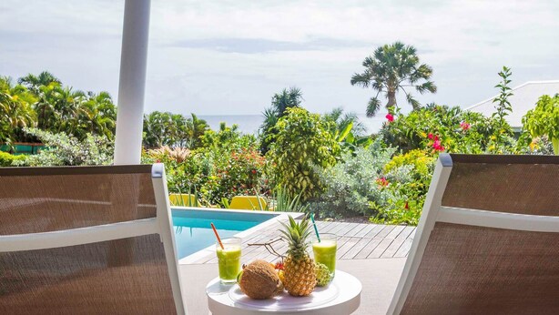 Un espace aménagé au bord de la piscine, avec deux chaises longues, une petite table sur laquelle se trouvent un ananas et des boissons, et une vue sur l’océan et la végétation tropicale.