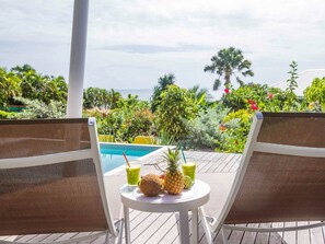 A poolside area with two lounge chairs, a small table with a pineapple and drinks, and a view of the ocean and tropical vegetation.