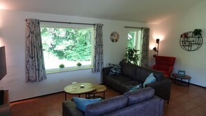 Ein Wohnzimmer mit Sofa, Sessel und Couchtisch. Zwei Fenster mit Vorhängen, eine Uhr an der Wand und eine kleine Pflanze an der Fensterbank.