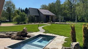 Una piscina con borde de piedra, cubierta de madera y una casa con techo inclinado en un jardín ajardinado.