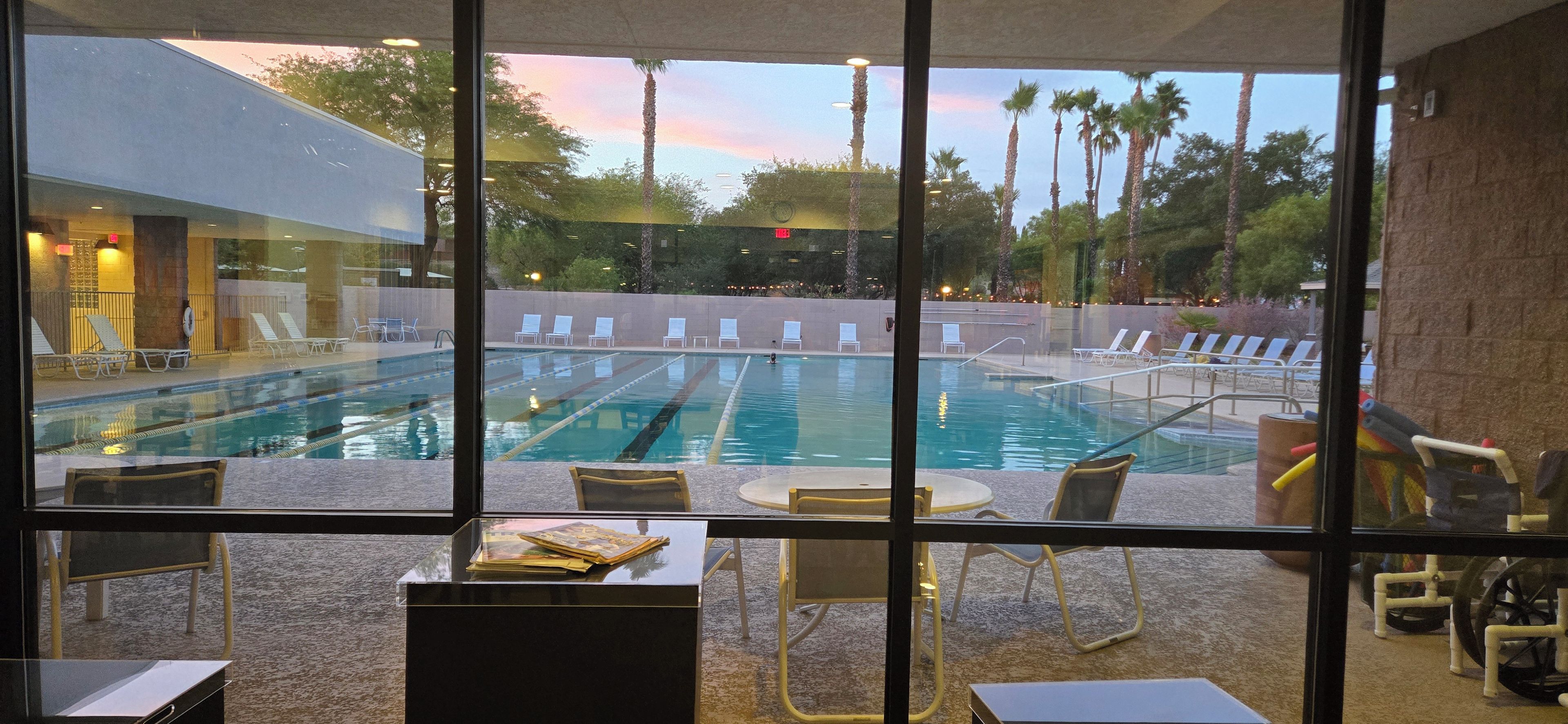 A pool area with lounge chairs and a table, viewed through a window with a view of palm trees and a sunset.