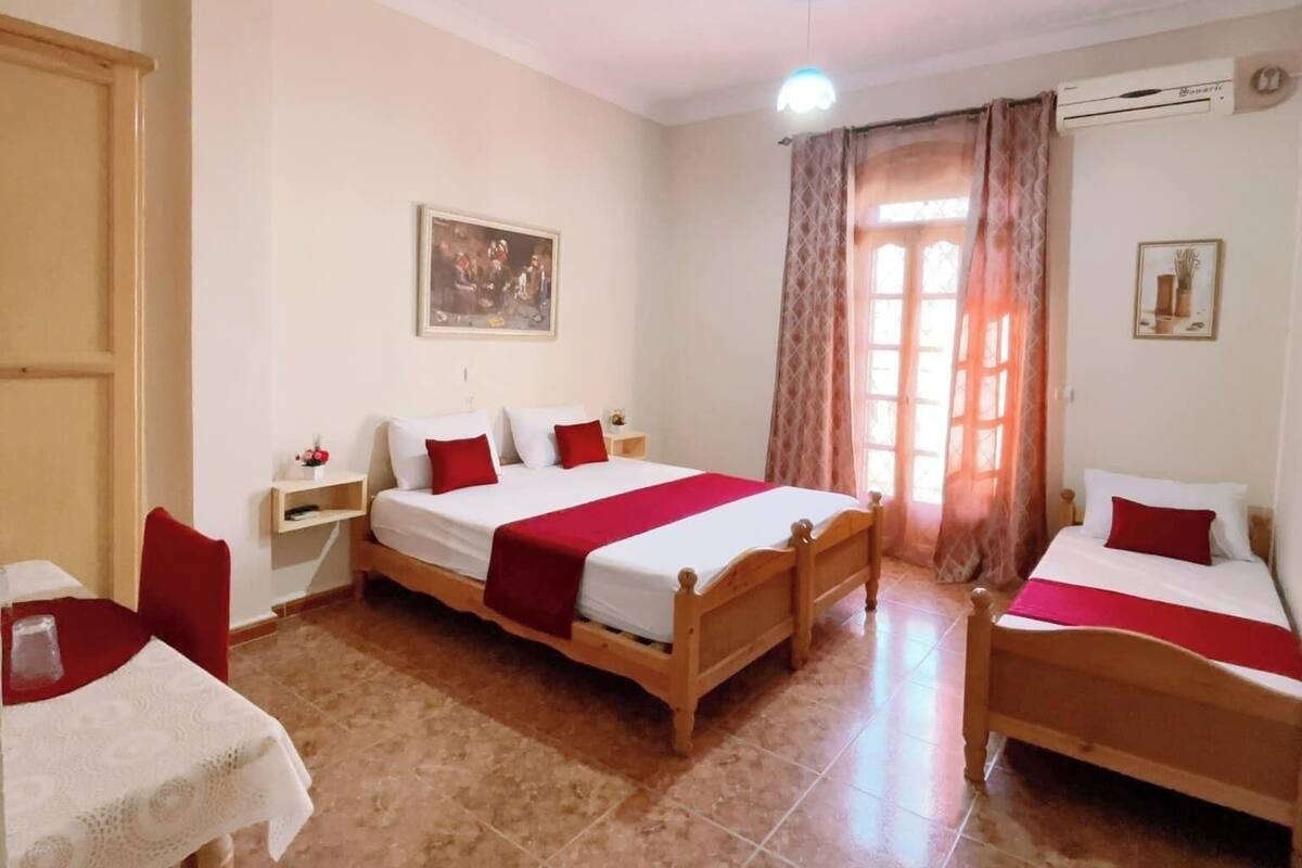 A hotel room with two wooden beds, a small table with a white tablecloth, a red chair, a nightstand, and a wall-mounted air conditioning unit.