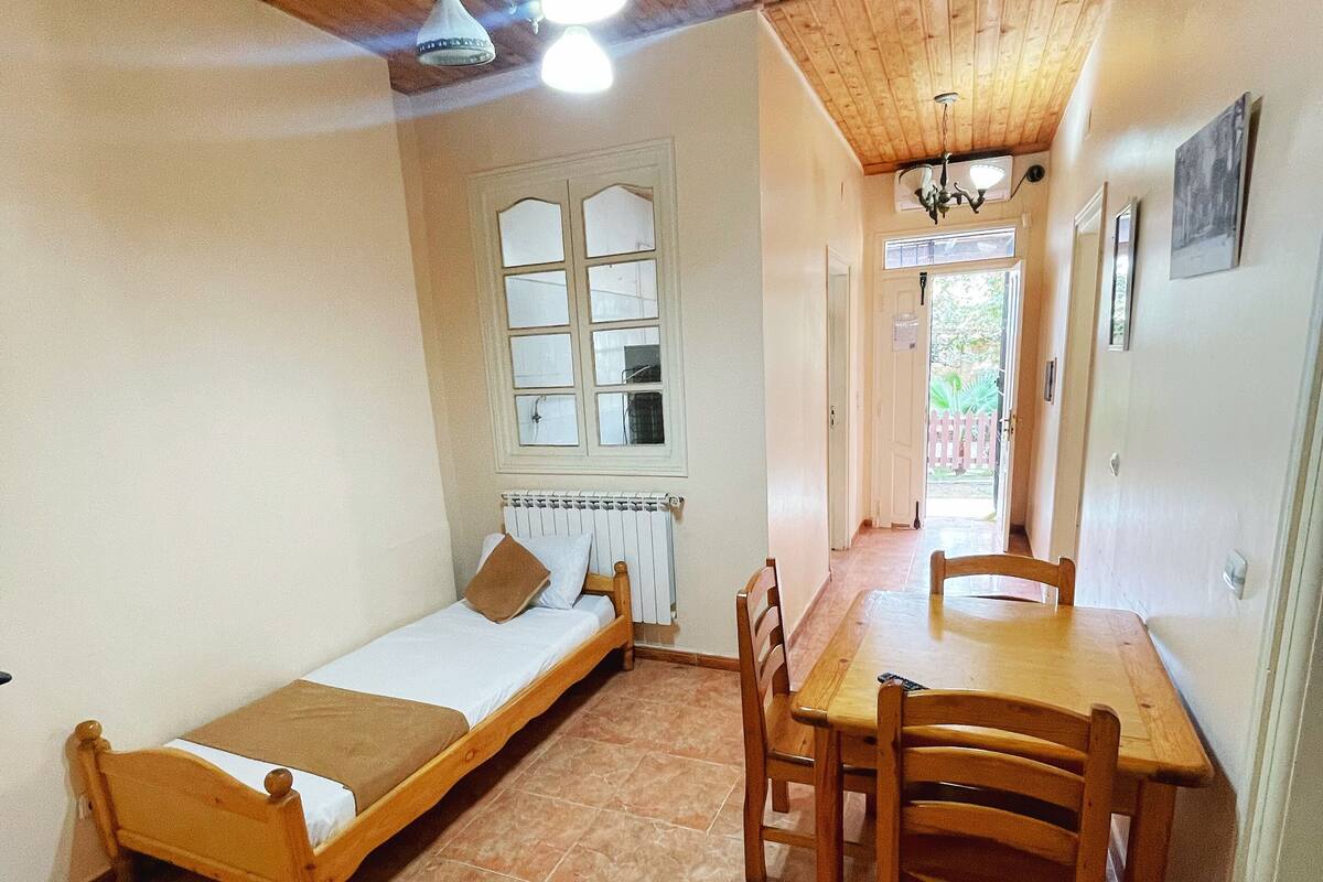 A small, cozy room with a single bed, a wooden table, and chairs. There is a window with a view of the outside, and a radiator under the window.