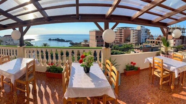 Une terrasse extérieure avec des tables et des chaises, une vue sur la mer et des bâtiments en arrière-plan.