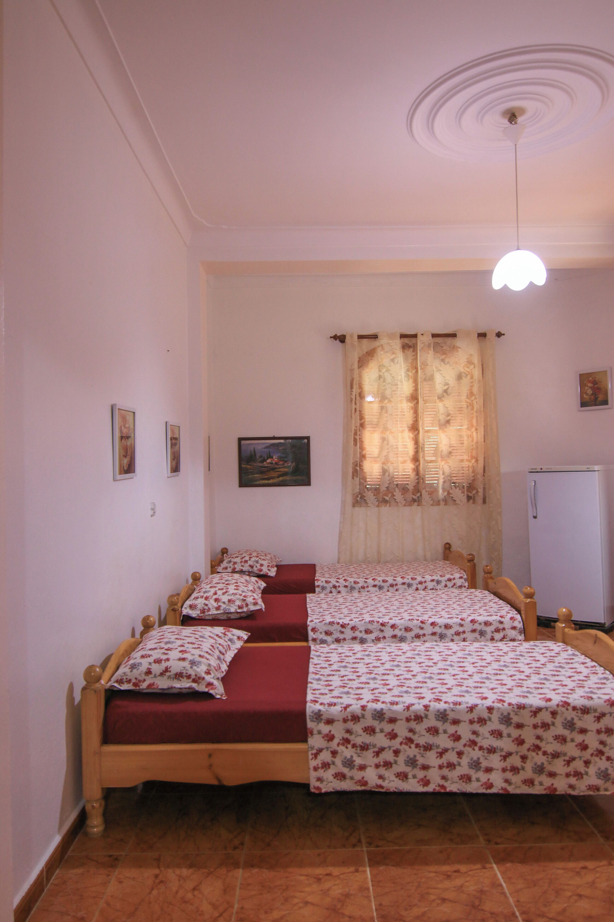 A bedroom with two single beds, a ceiling fan, a window with curtains, a refrigerator, and a wall-mounted picture.