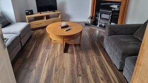 A living room with a round wooden coffee table, a grey sofa, a grey armchair, a wooden TV stand, and a fireplace. - 3-beds, Sleep 7 With Garden & Private Parking (Carrick-on-Shannon)